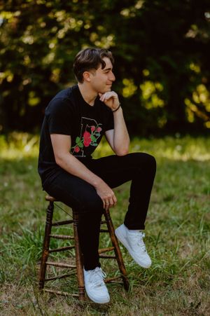 Person sitting on a wooden chair in a grassy field wearing black clothing and white shoes.
