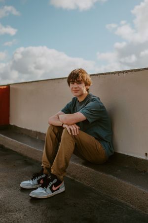 Person sitting on rooftop ledge wearing casual clothes and sneakers against cloudy sky.