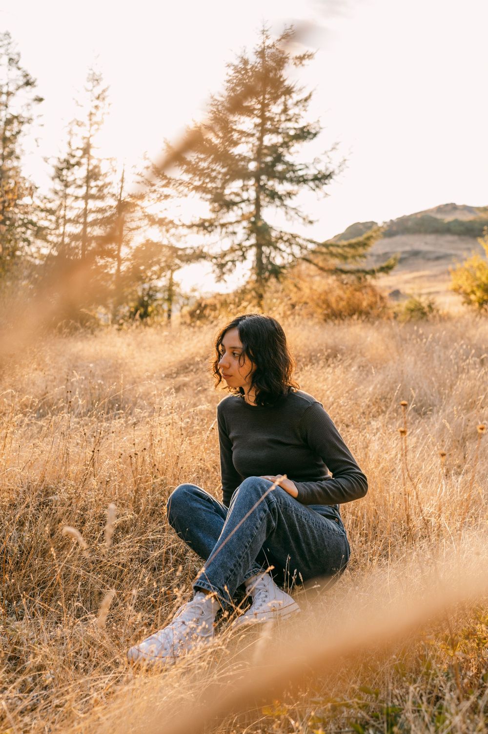 Person in dark clothing sitting in golden grass during sunset with mountains in background.