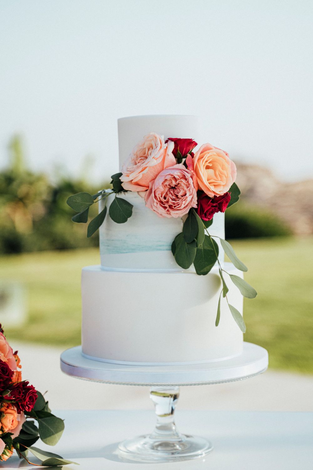 Elegant two-tier white wedding cake decorated with pink and burgundy roses on a cake stand.