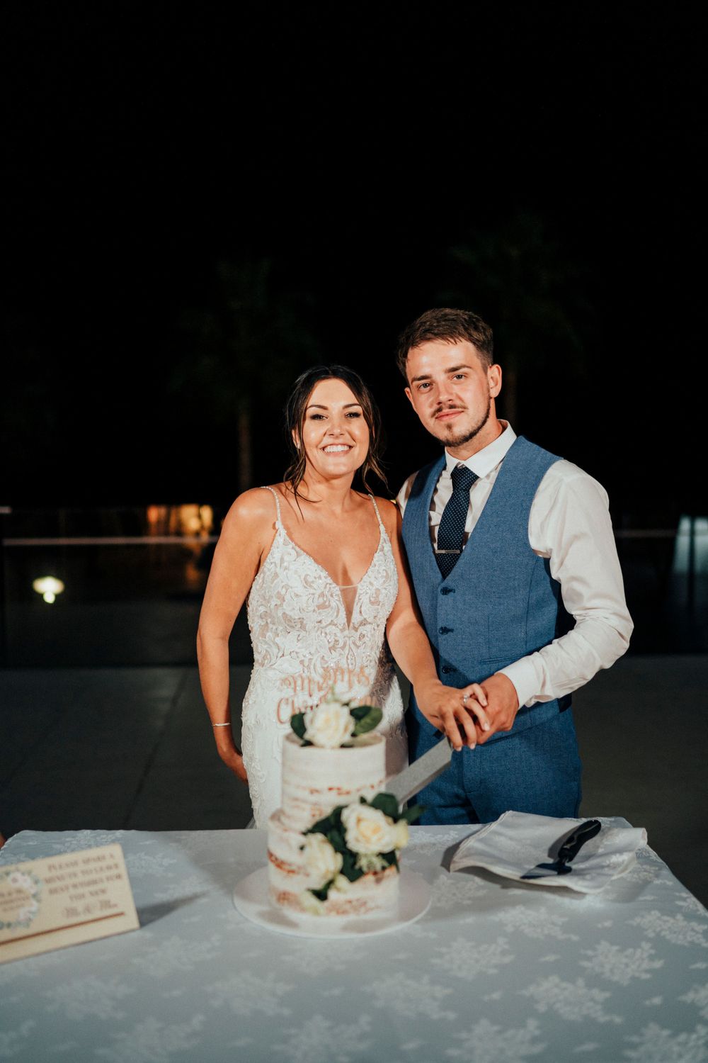 A couple stands behind their wedding cake at an evening reception.