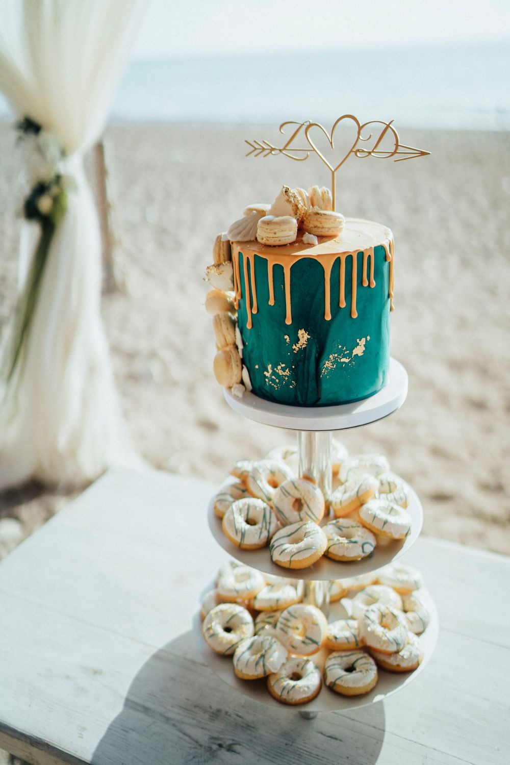 Teal and gold drip wedding cake with macarons on tiered dessert stand at beach wedding.