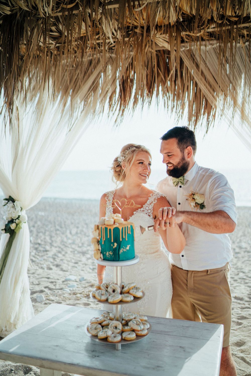 A couple cuts into a teal and white wedding cake under a thatched beach pavilion.
