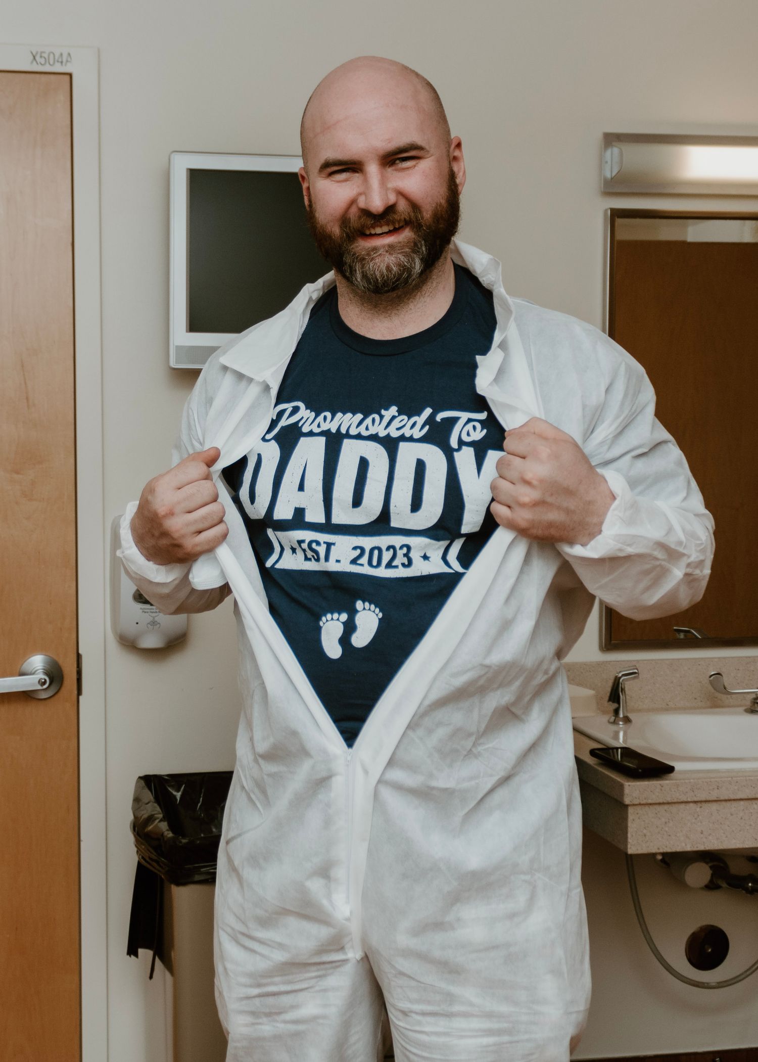 Person in white protective suit showing off 'Promoted to Daddy 2023' t-shirt in medical setting.