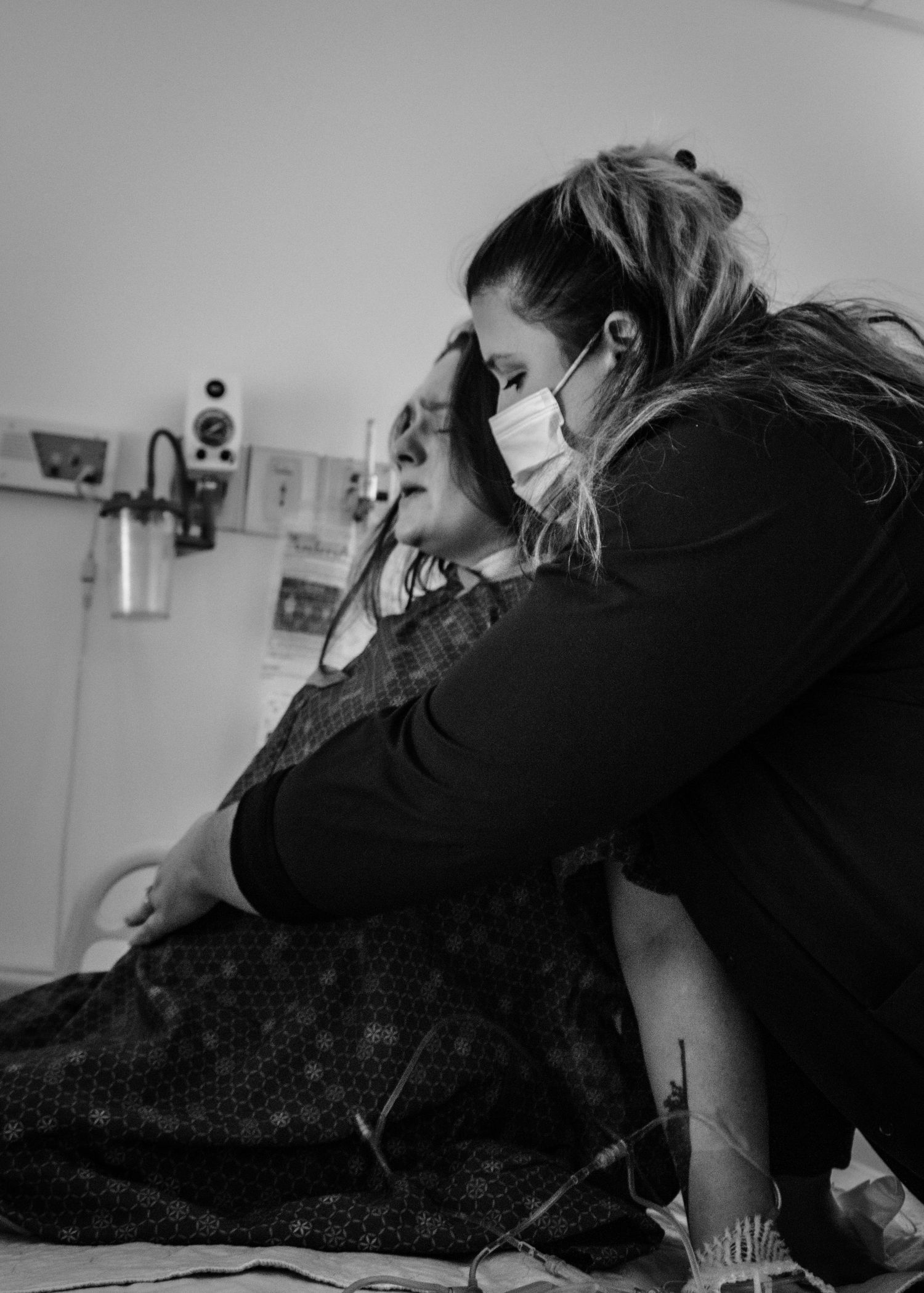 Healthcare worker wearing protective mask comforts patient in hospital room during emotional moment.