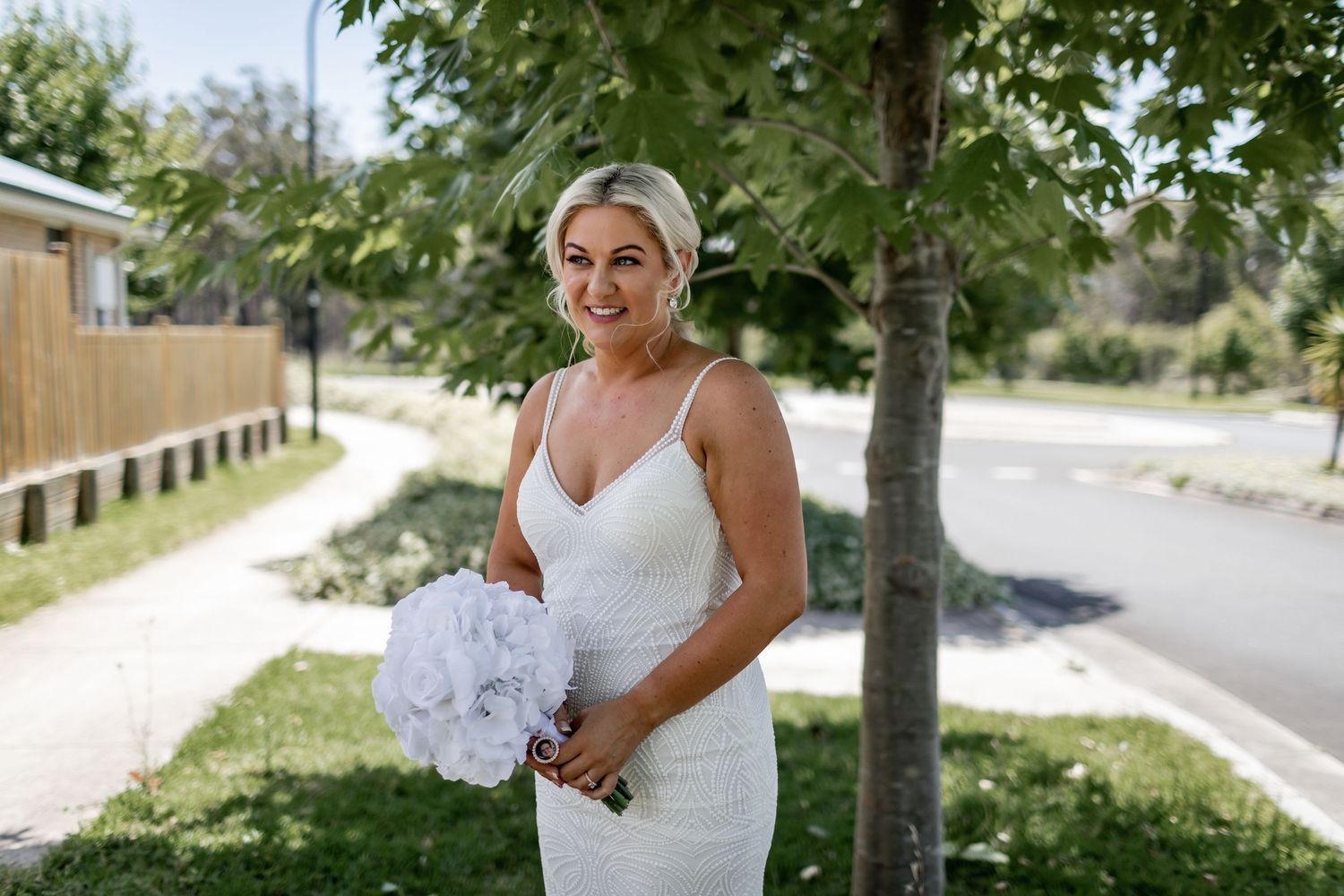 Bride in elegant white dress posing outdoors with bouquet on sunny day.