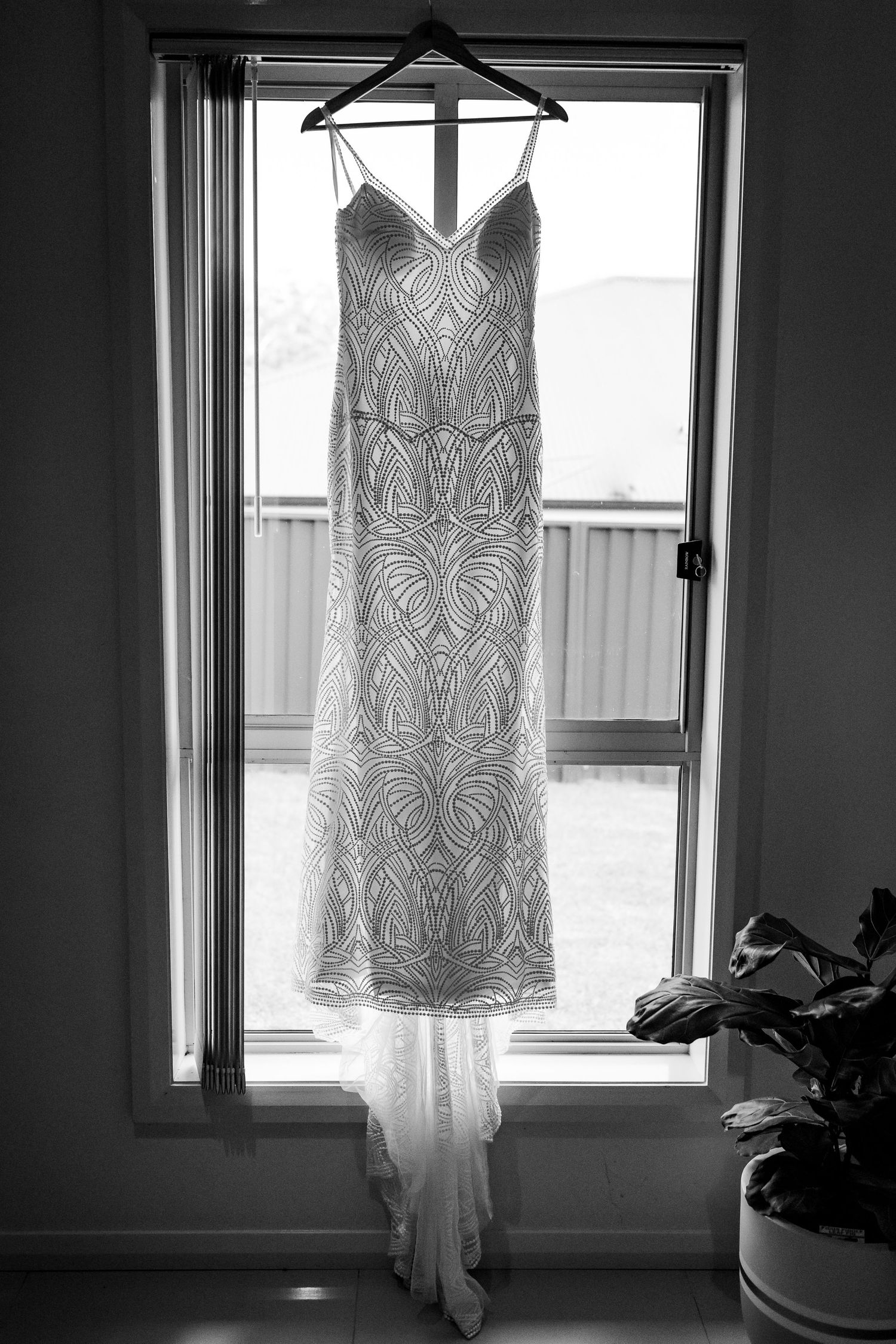 Elegant white lace wedding dress with intricate patterns hanging in window silhouette against natural light.