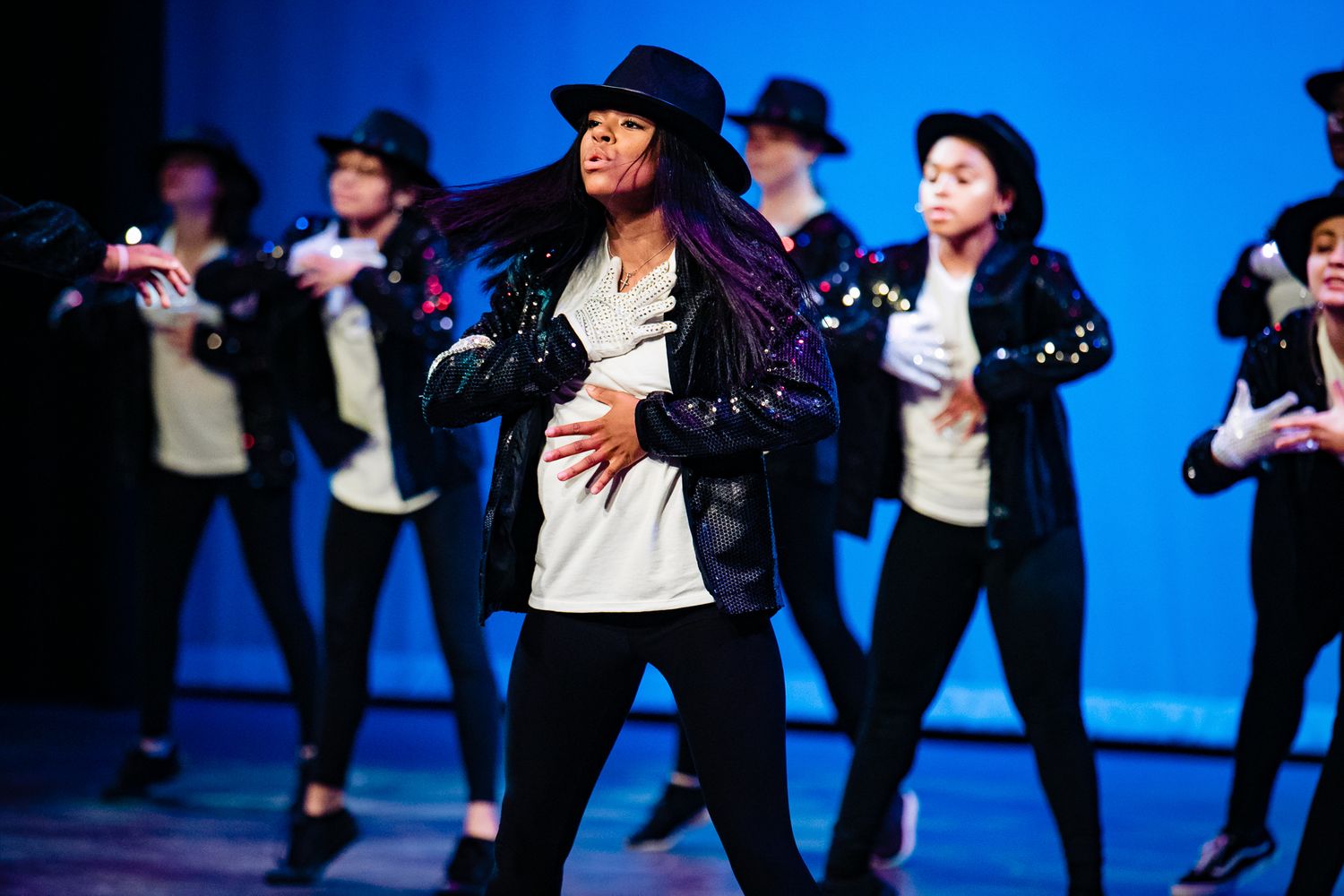 Dancers performing choreographed routine in black costumes against blue background.