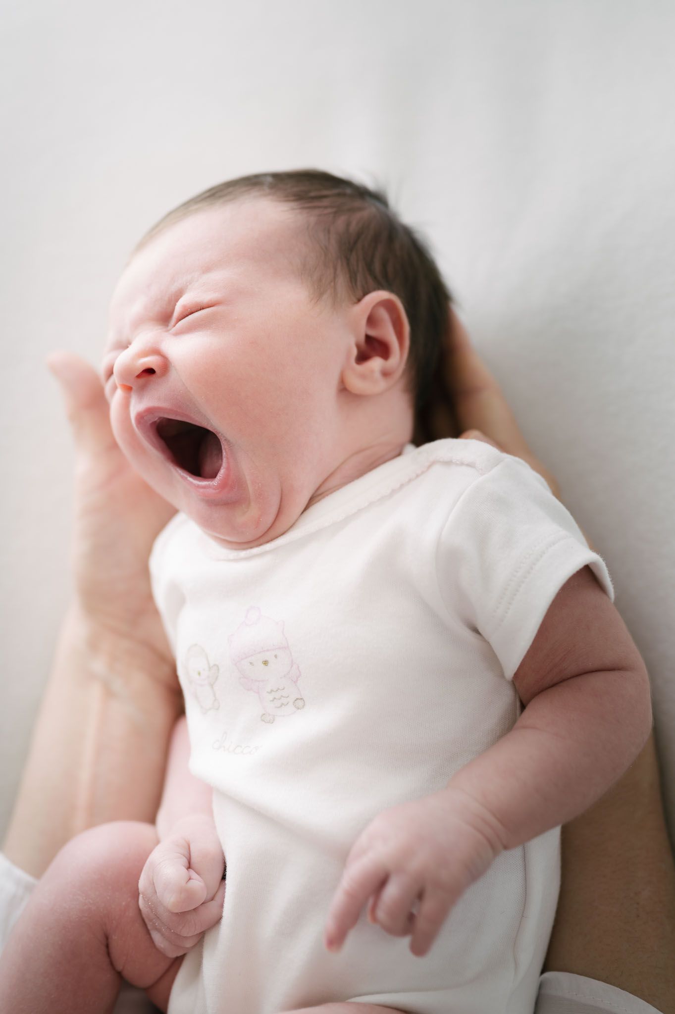 Candid photo of yawning newborn baby in white onesie against light background.