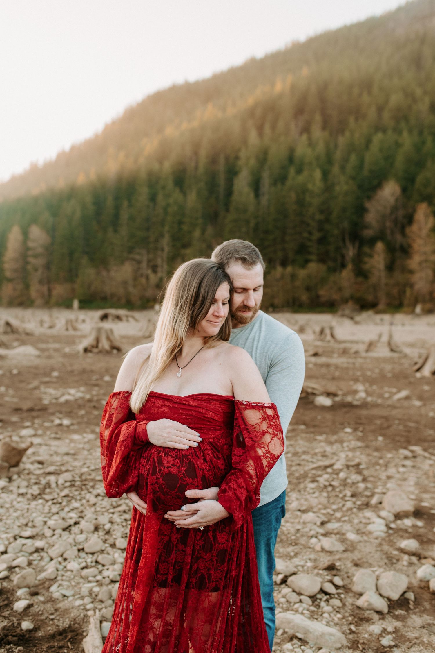Romantic maternity photos by rocky riverbed with couple in red dress and casual wear at sunset.