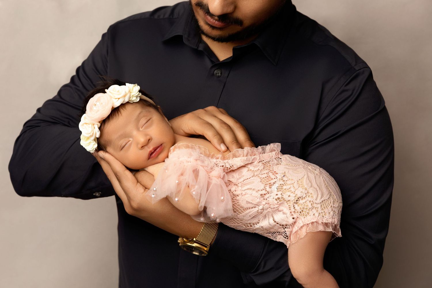 Father in black shirt gently cradles sleeping newborn baby girl in pink lace dress with floral headband.