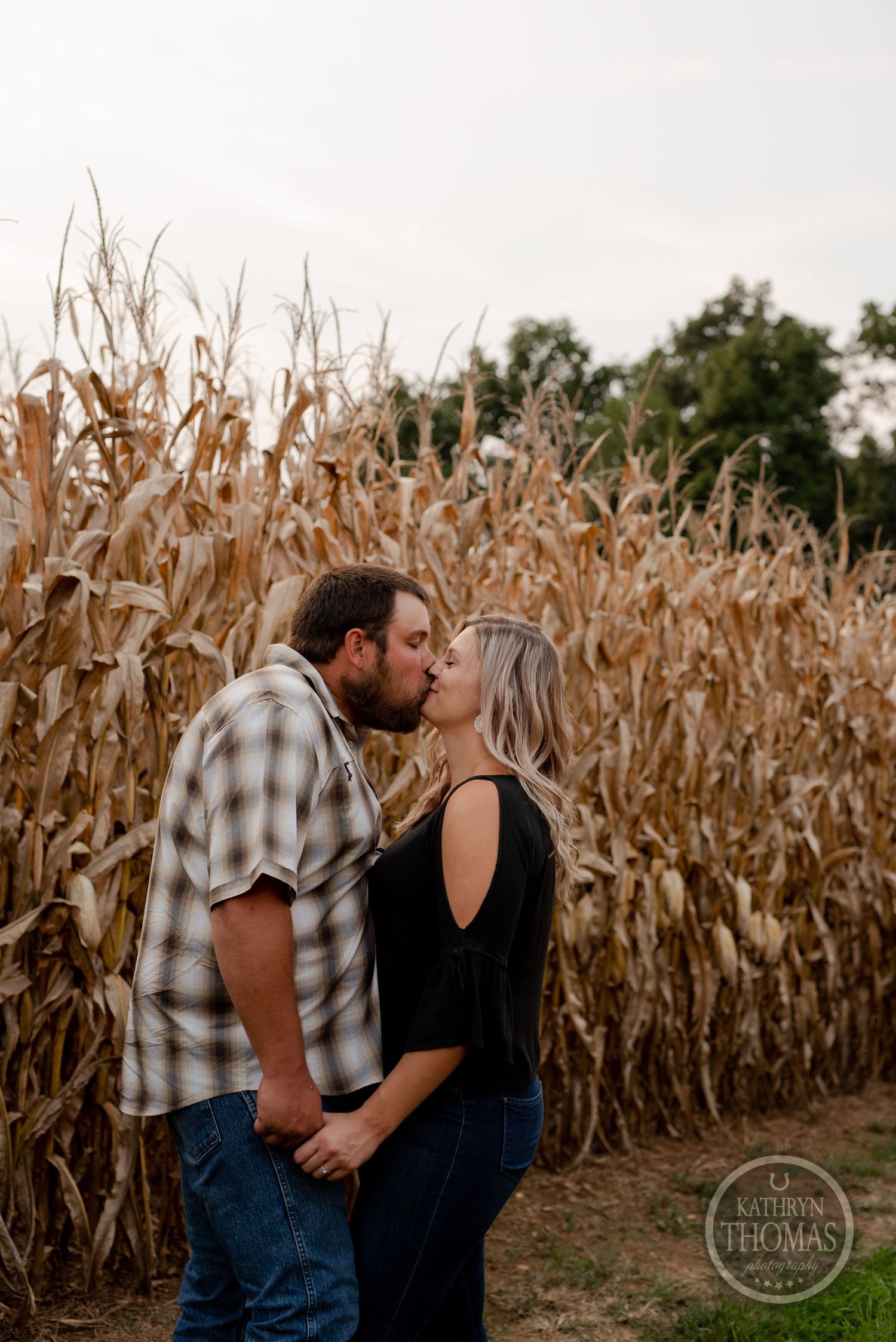 Intimate moment between couple embracing in front of tall corn stalks at sunset.