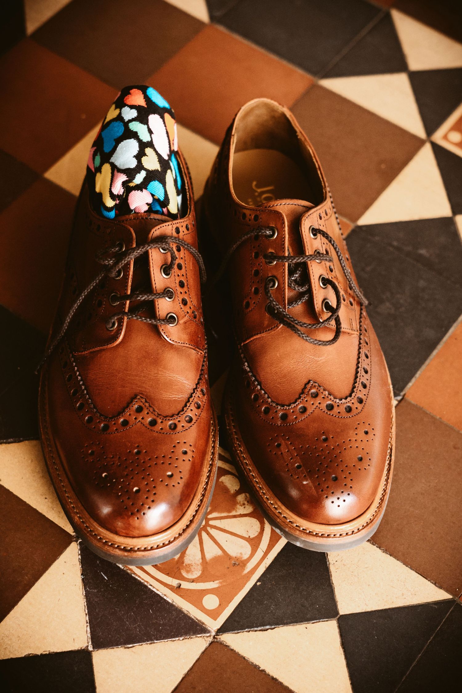 Brown leather brogue dress shoes with colorful socks on a geometric patterned floor.