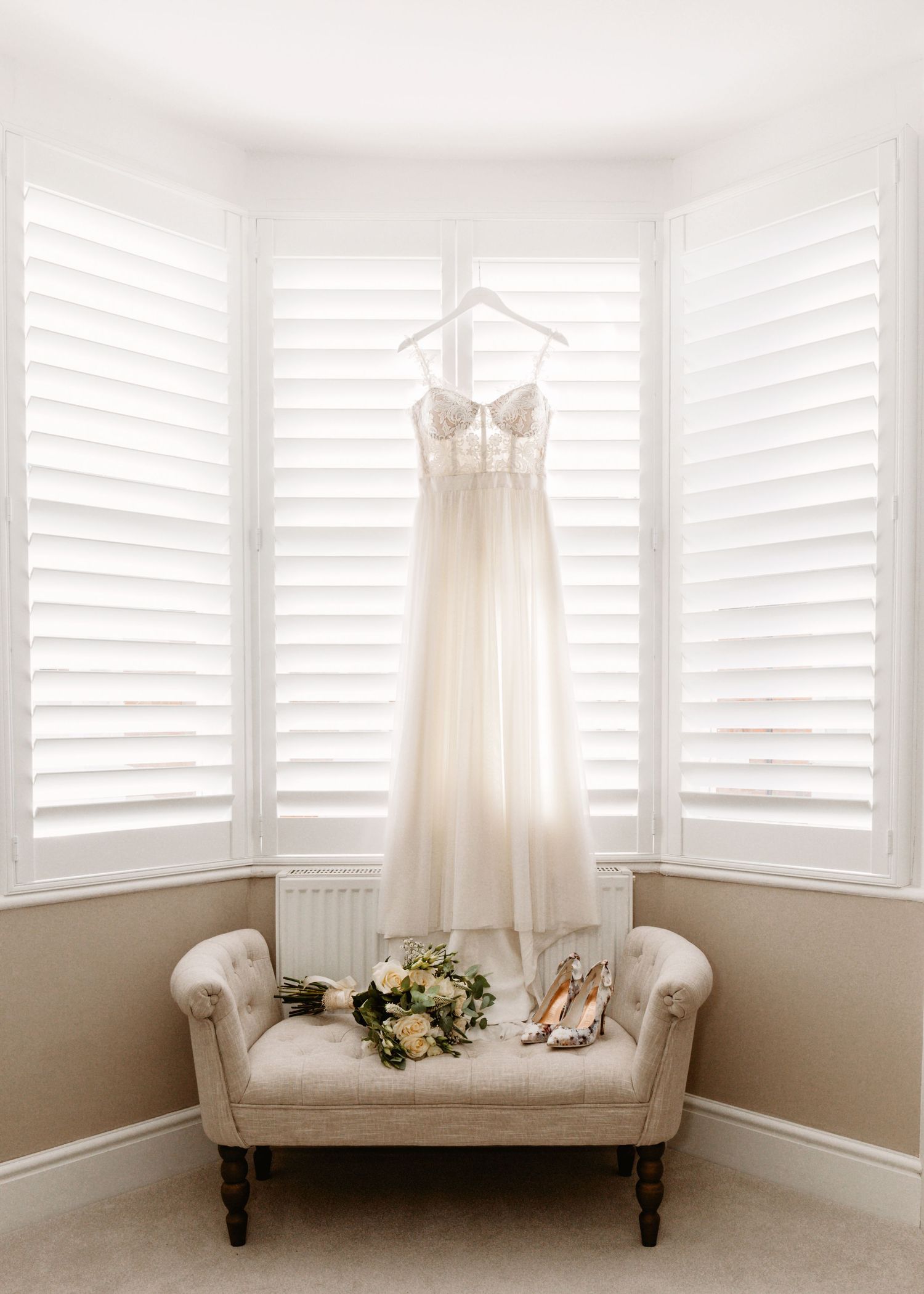 White wedding dress hanging on a hanger above a beige settee with floral bouquet and shoes.
