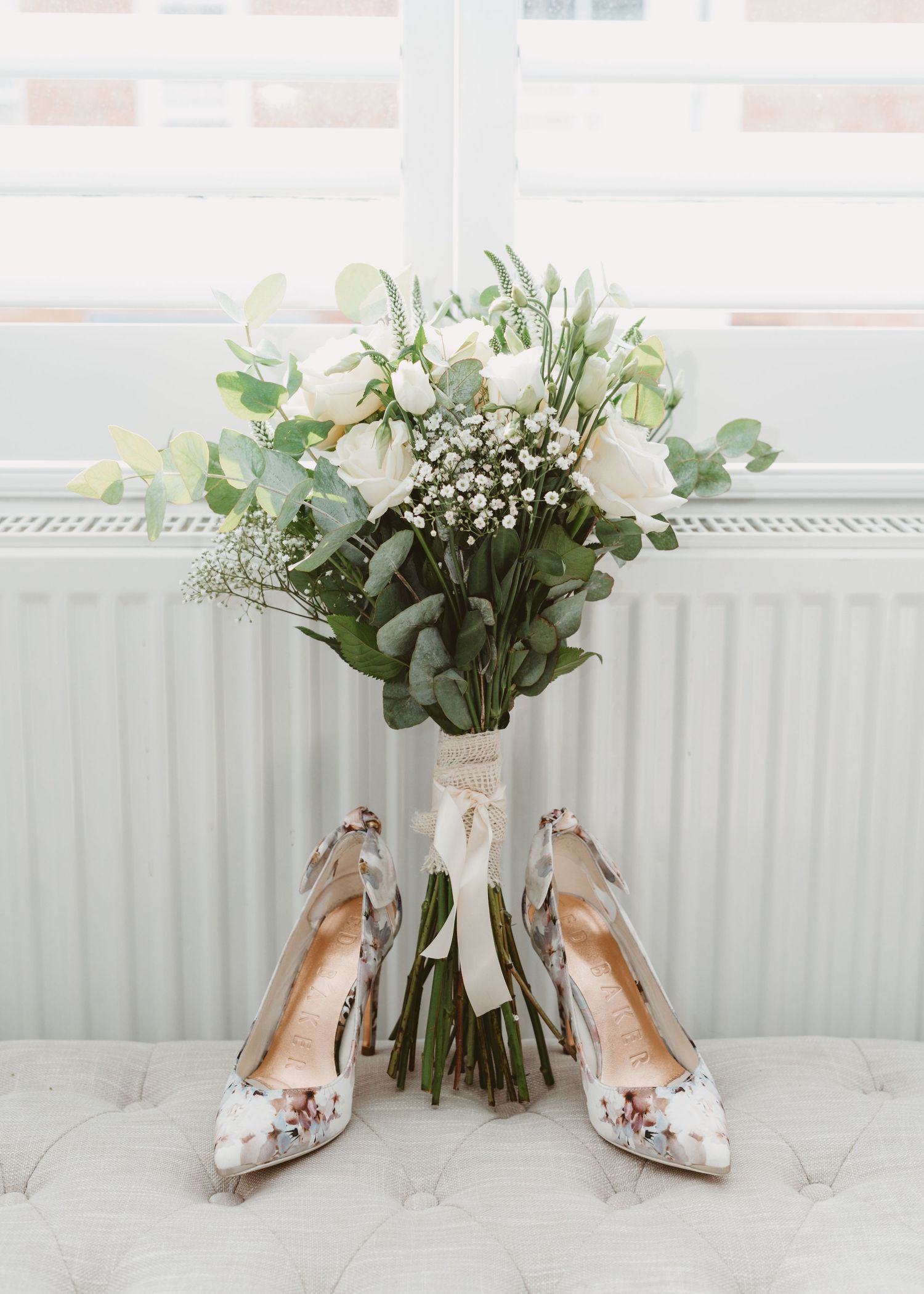Wedding bouquet of white roses and eucalyptus with matching floral high heel shoes arranged on a windowsill.