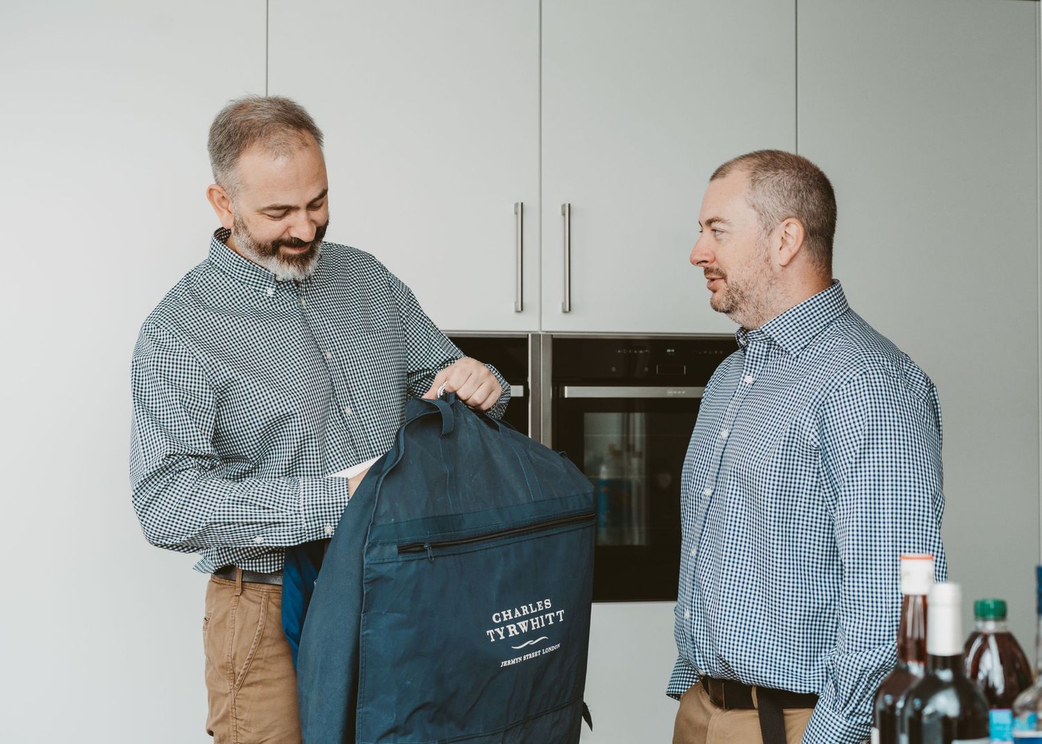 Two men in matching blue checkered shirts sharing a laugh while holding a navy blue bag.