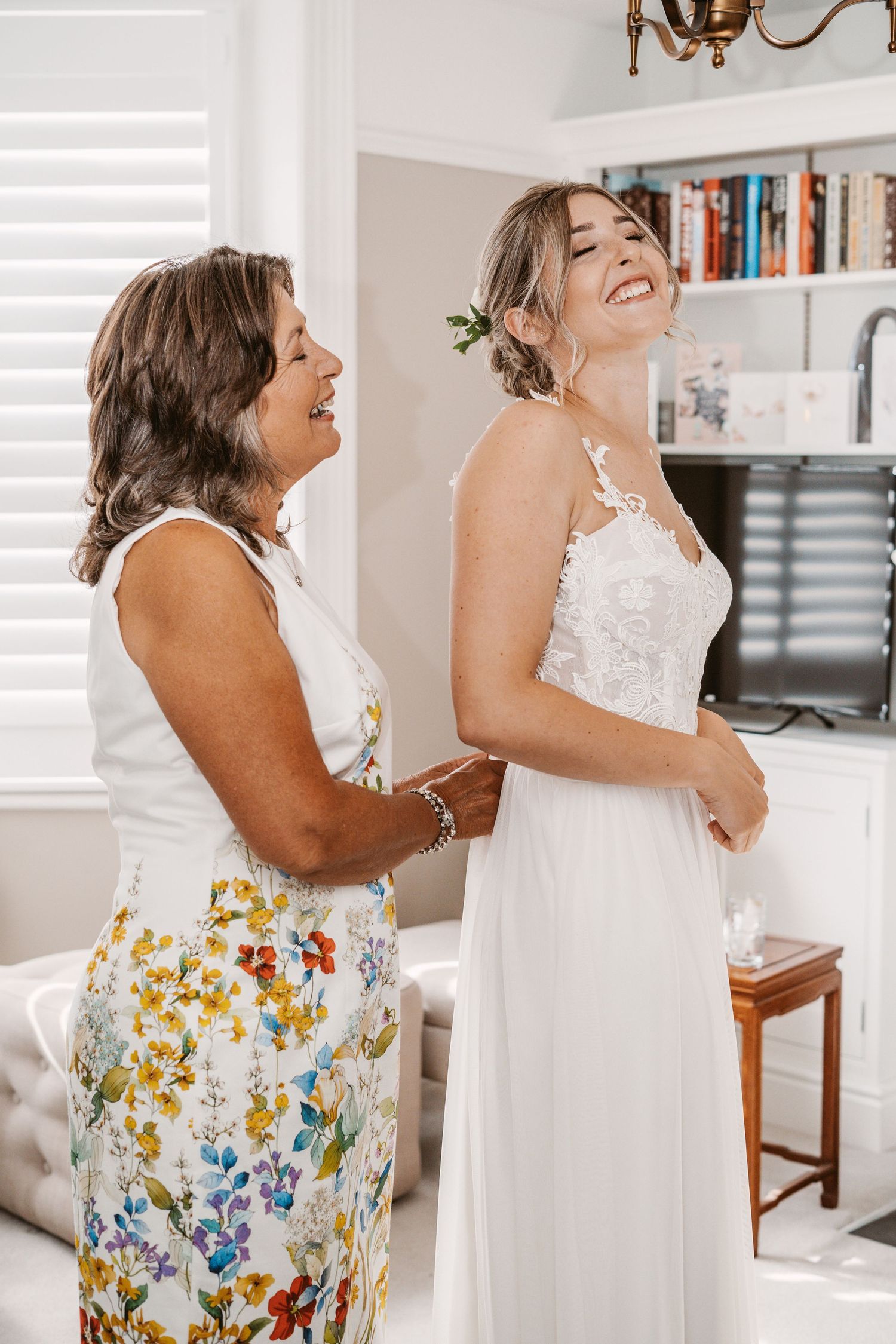 Wedding dress fitting moment captured in bright interior with white shutters and bookshelves.