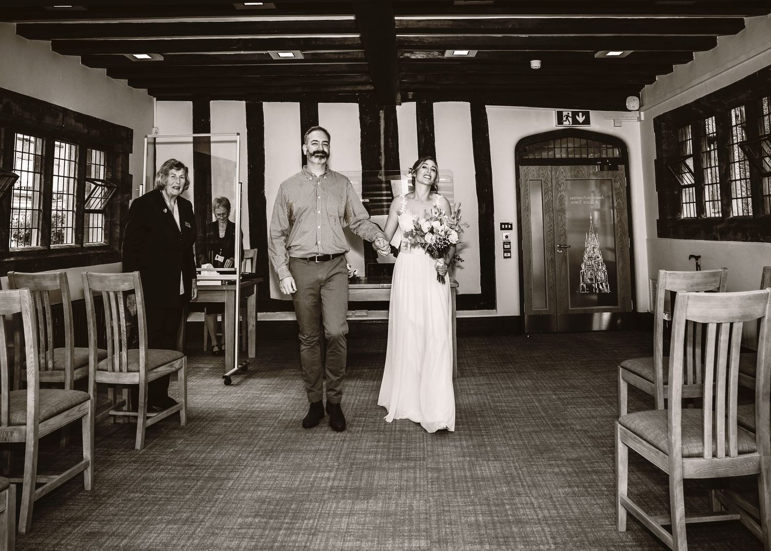 Black and white photograph of wedding ceremony in historic building with wooden chairs.