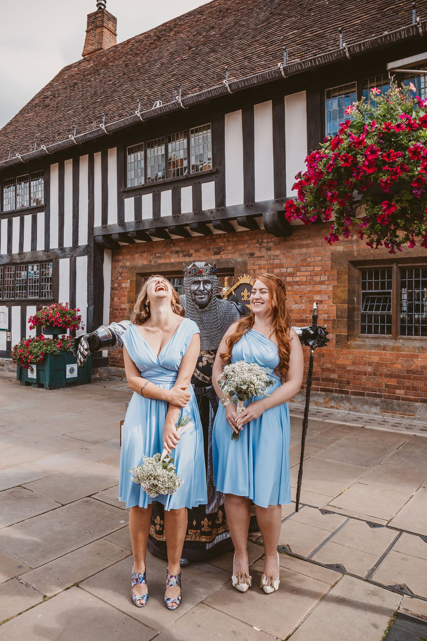 Two bridesmaids in light blue dresses posing next to a statue in front of a Tudor-style building.