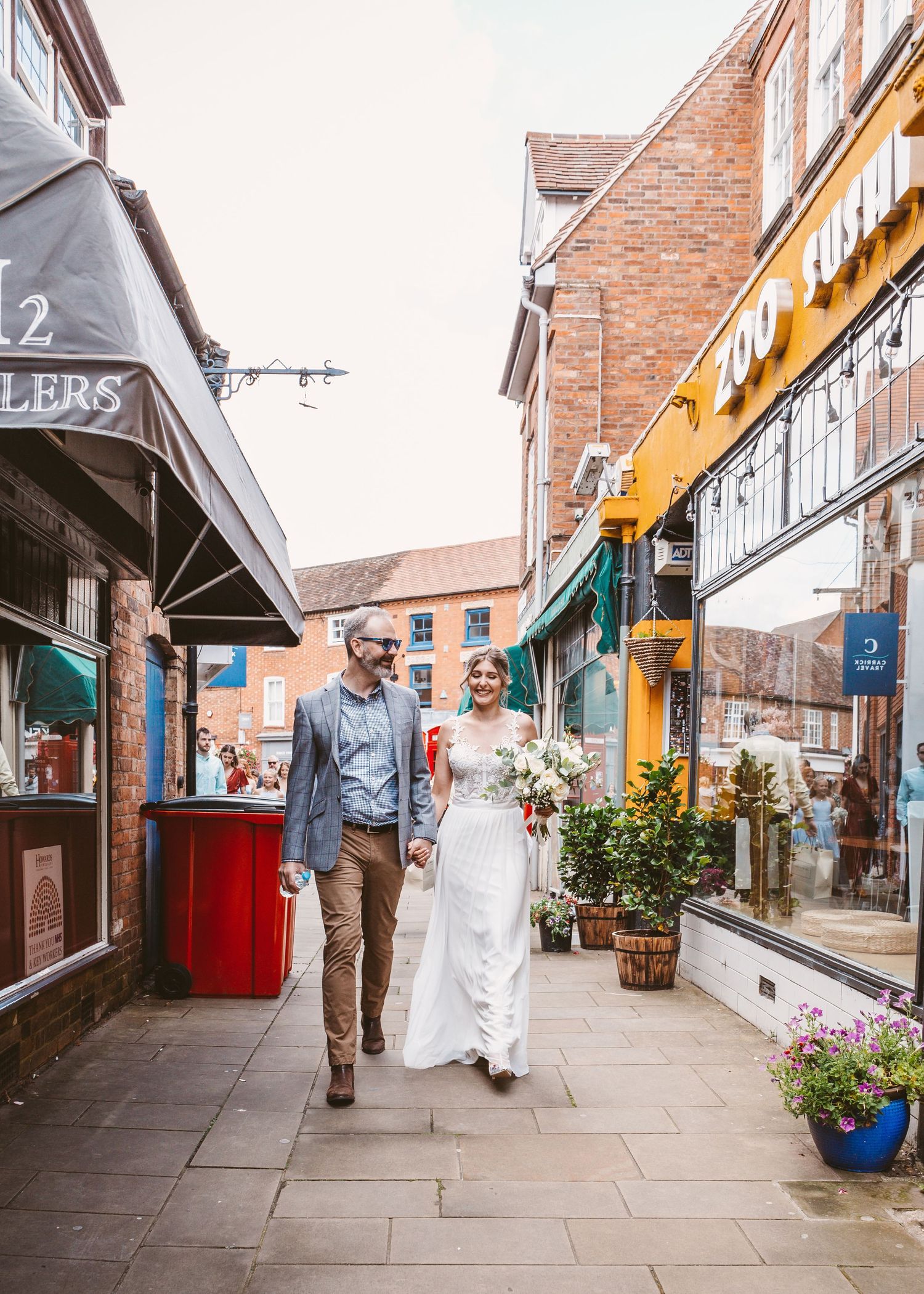 A couple walking along a quaint shopping street with brick buildings and flower planters.