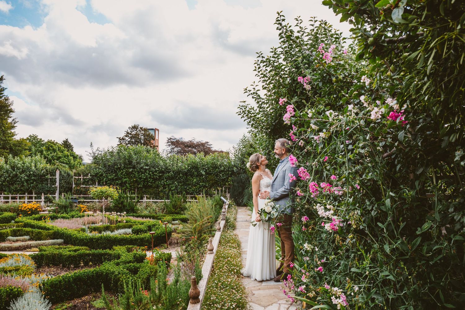 A couple standing in a beautiful formal garden with ornate hedges and pink flowering bushes.