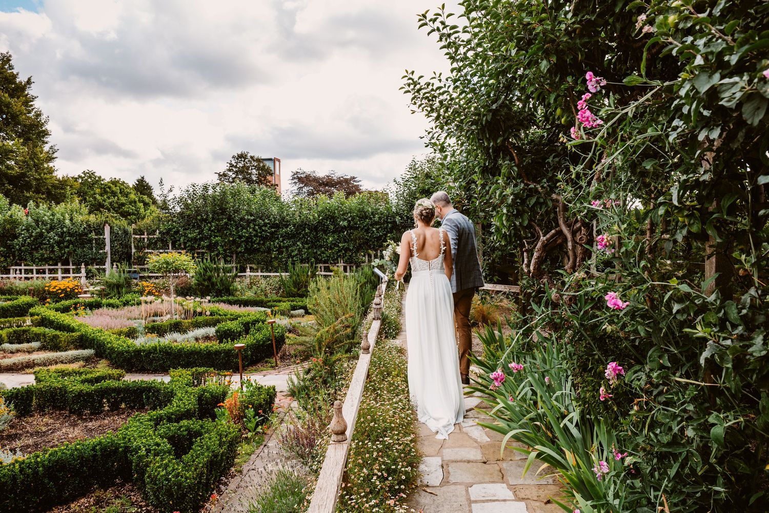 A white-dressed figure walks along a stone path in a formal garden with geometric hedges and pink flowers.