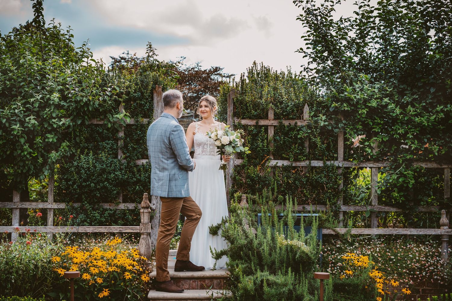 Couple standing on stone steps in a garden with vibrant yellow flowers and rustic wooden fencing.