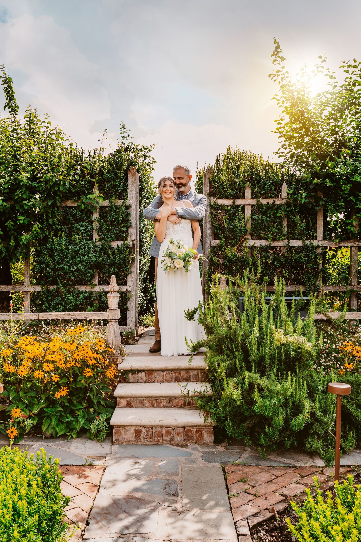 Wedding couple embracing on garden steps with golden sunlight streaming through trees.