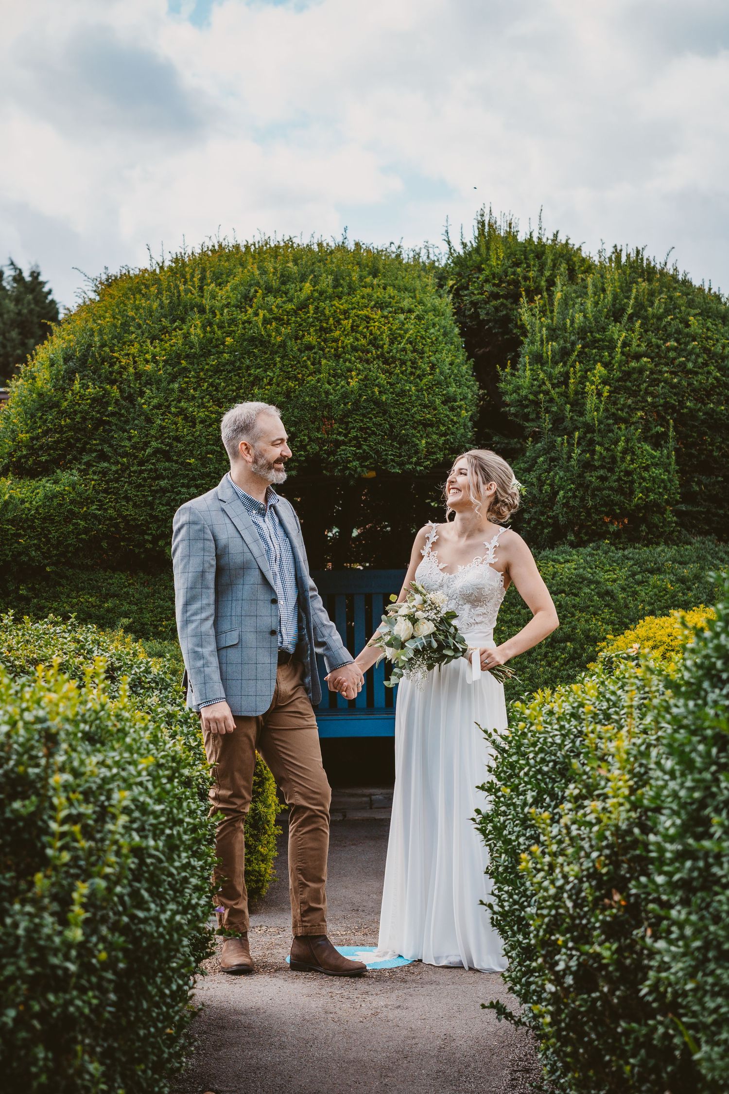 Wedding couple holding hands in a formal garden path surrounded by manicured hedges.