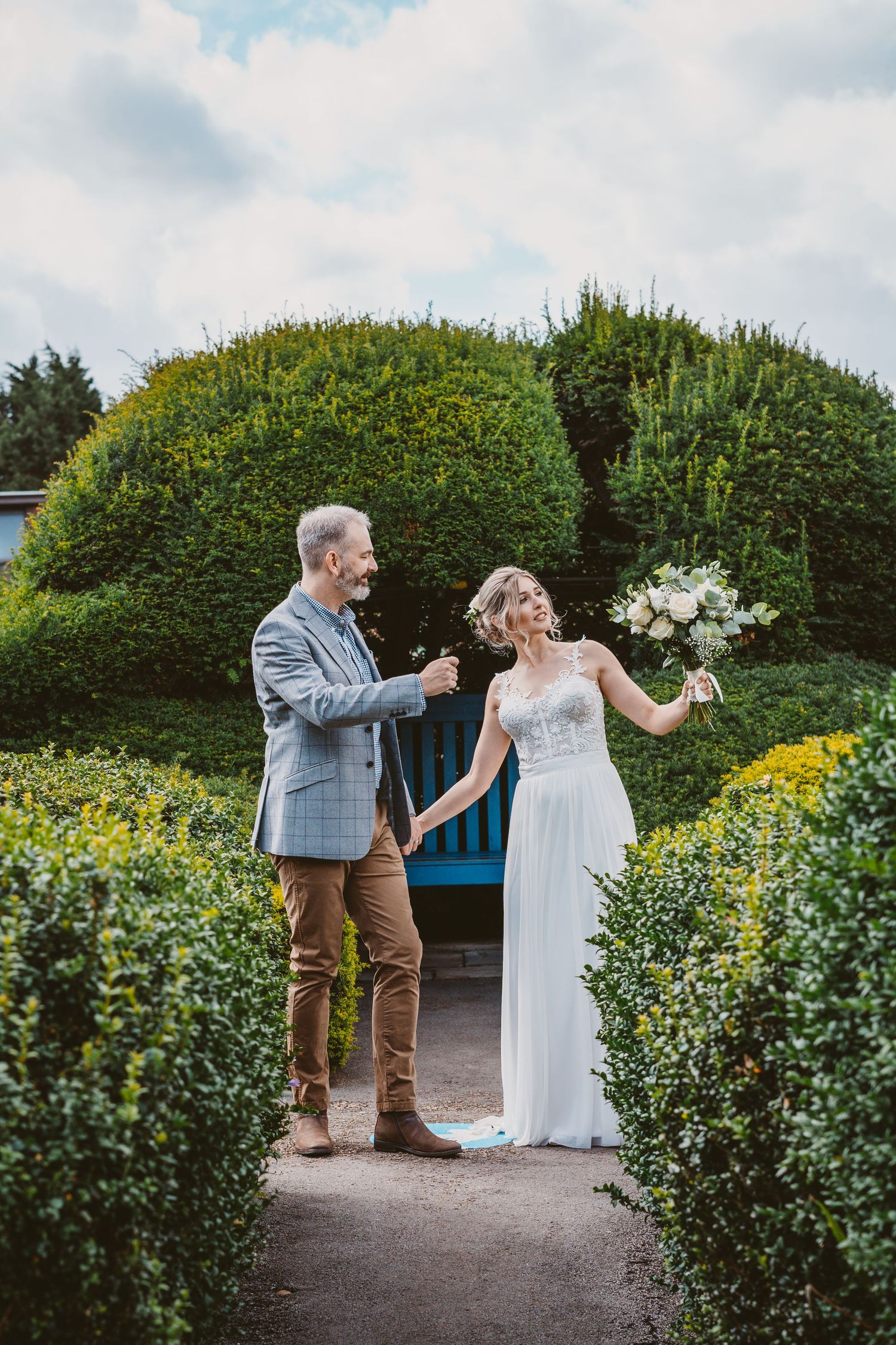 Joyful wedding couple dancing and laughing in a garden path between sculpted hedges.