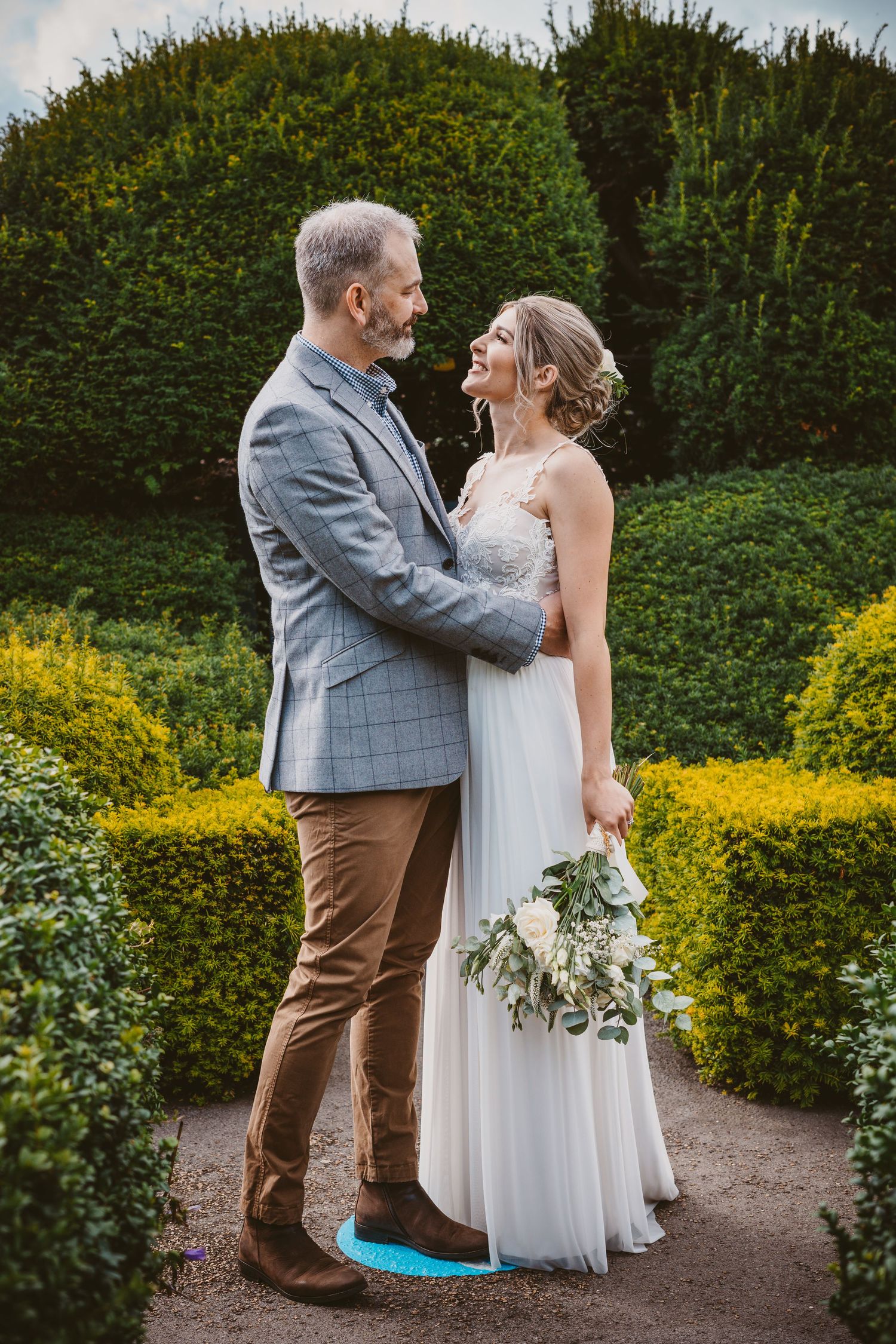 Intimate moment between wedding couple on garden path with manicured topiary backdrop.