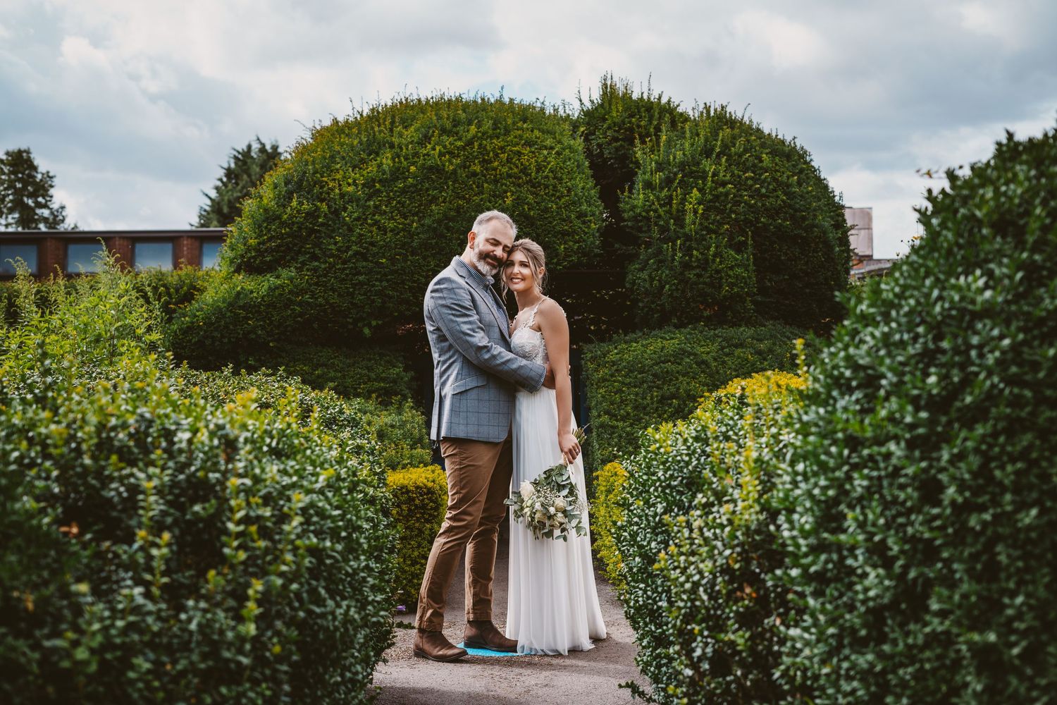 Wedding couple embracing on garden walkway with rounded topiary bush in background.
