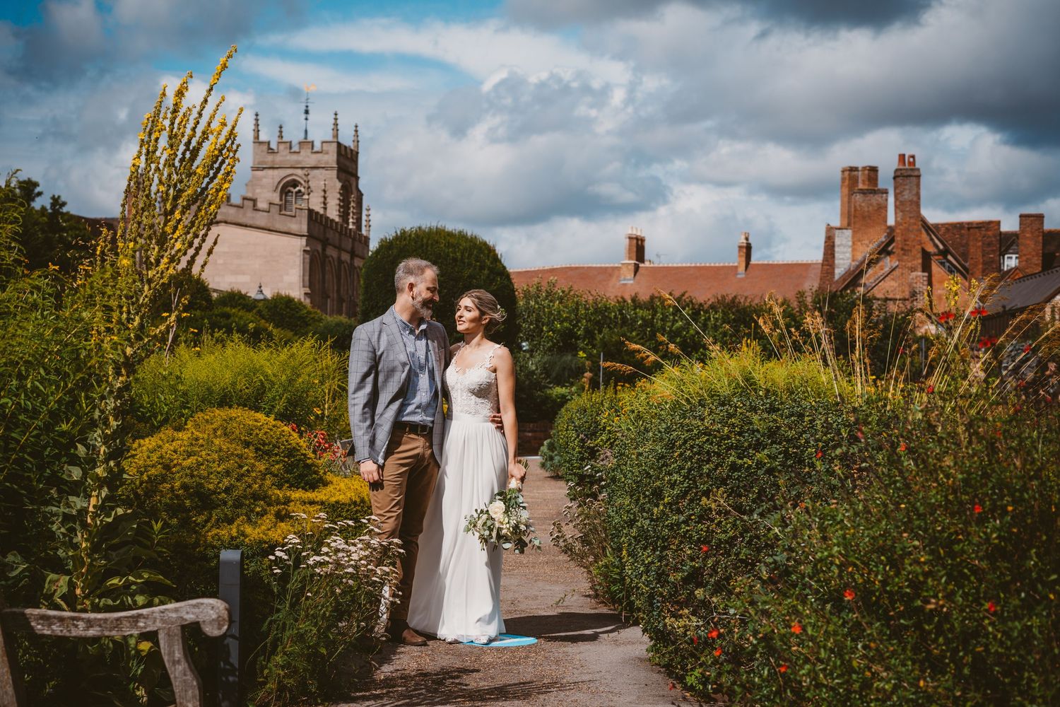 Wedding couple walking through English garden with historic church and buildings in background.