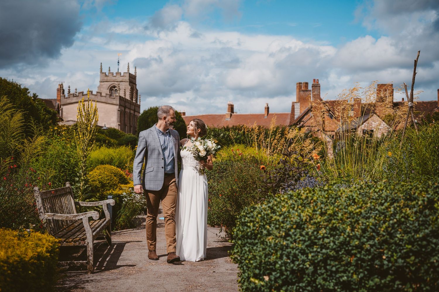 A romantic wedding couple walks along a garden path with a historic church and buildings in the background.