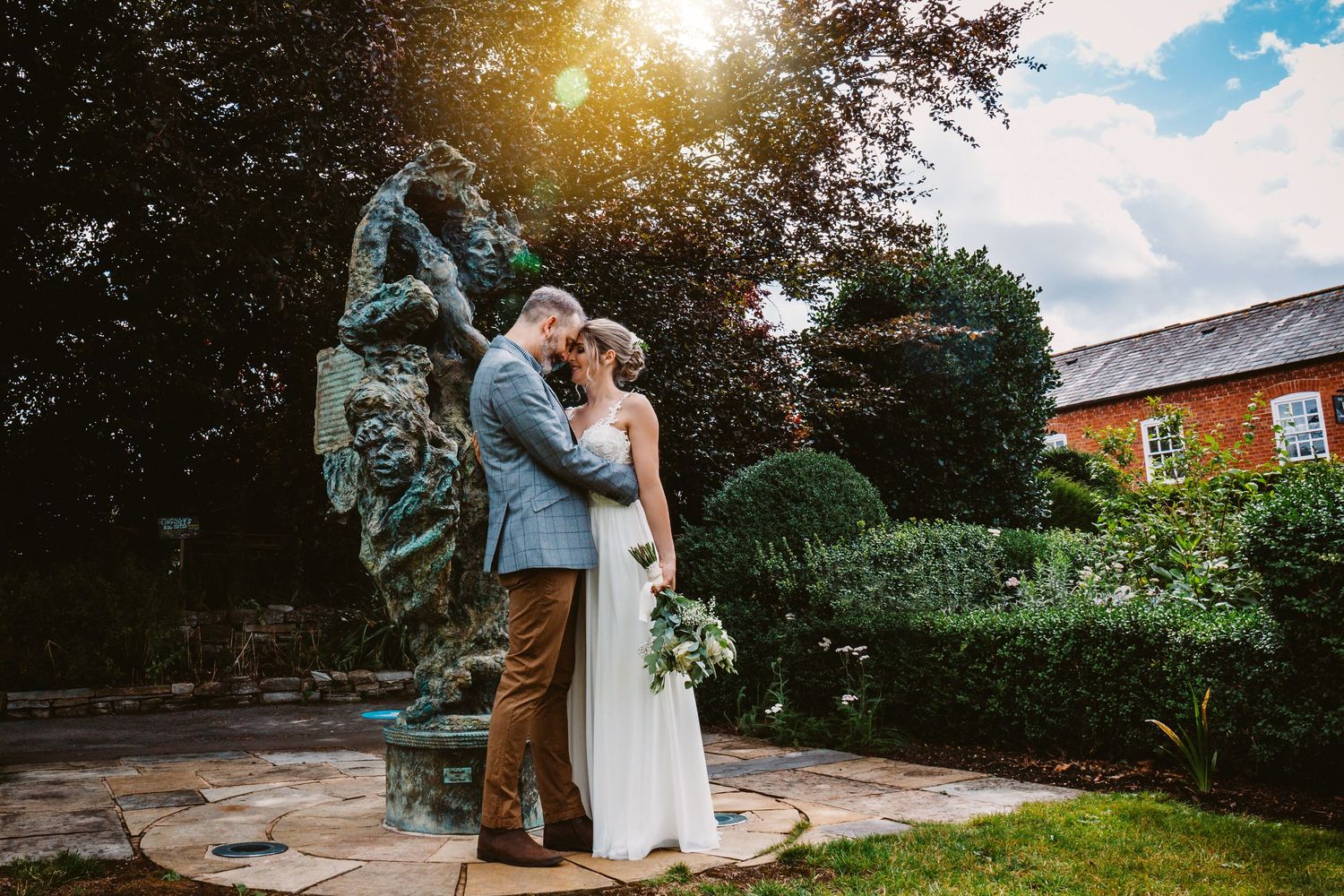A wedding couple embraces in a sunlit garden with ornate sculpture and red brick building behind them.