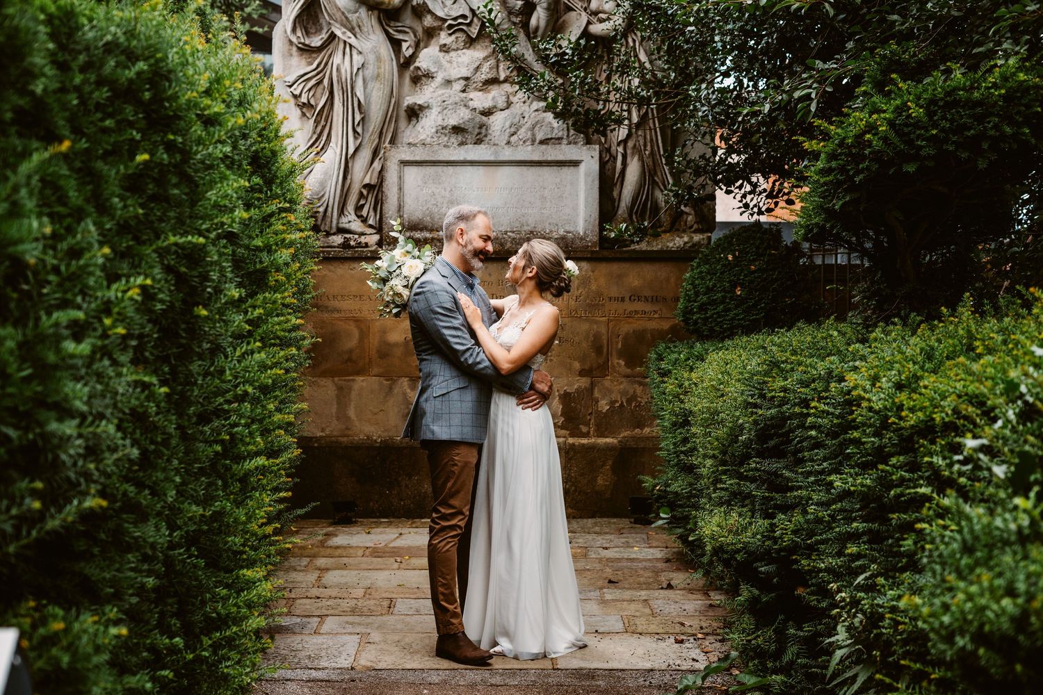A romantic wedding embrace in a garden setting with ornate stone sculptures and greenery.