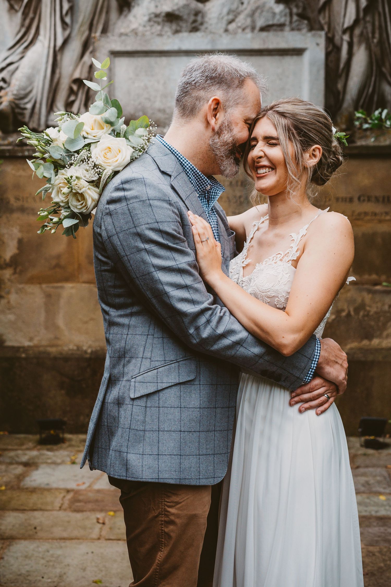 A tender moment between newlyweds with elegant white bouquet against stone wall background.