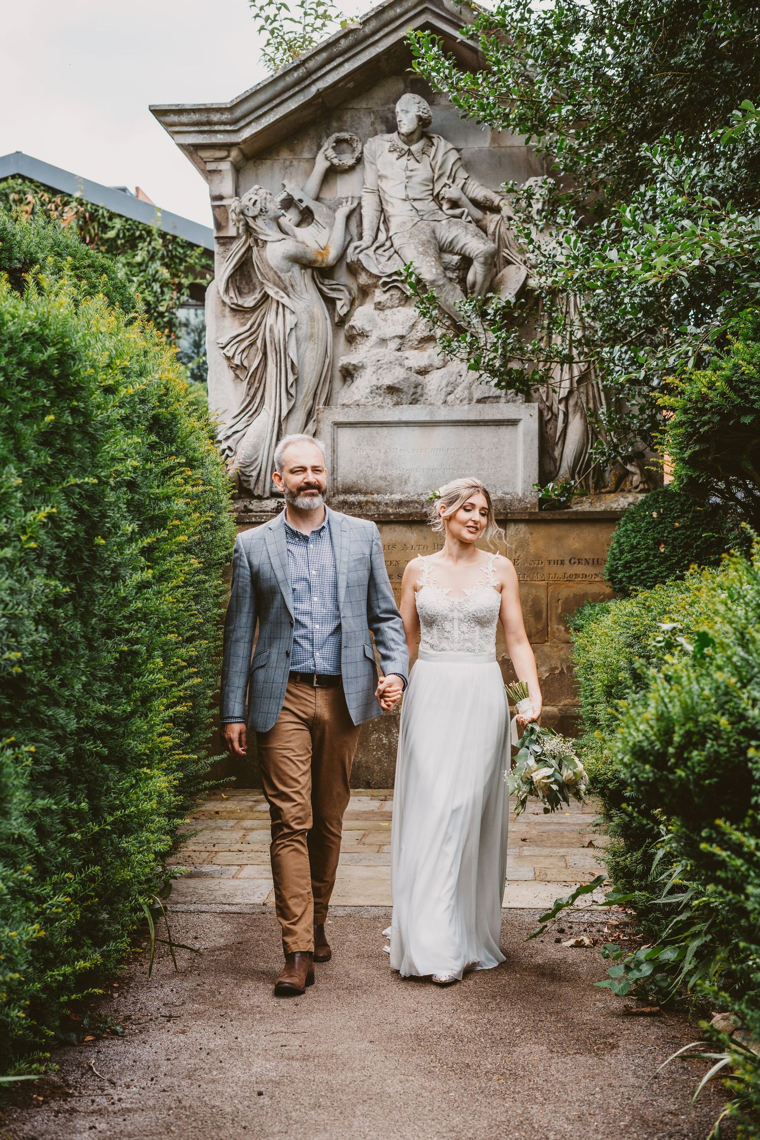 Newlyweds walking down garden path with ornate classical stone relief sculpture behind them.