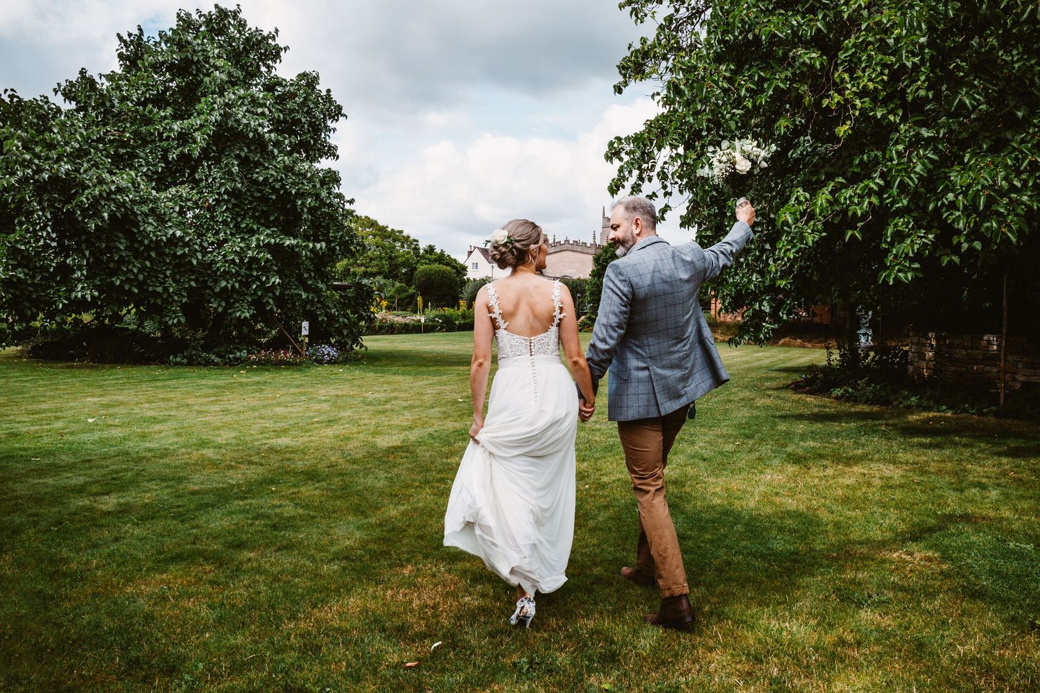 Joyful newlyweds walking hand in hand across a green lawn on their wedding day.