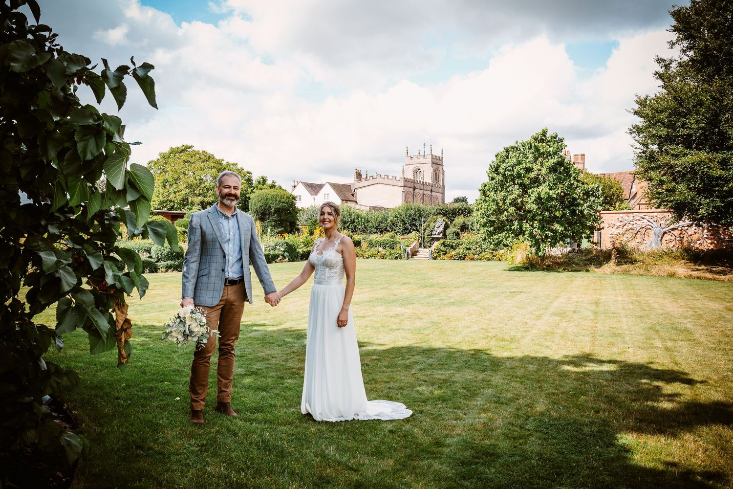 Wedding couple poses on lawn with historic church spires visible in background.