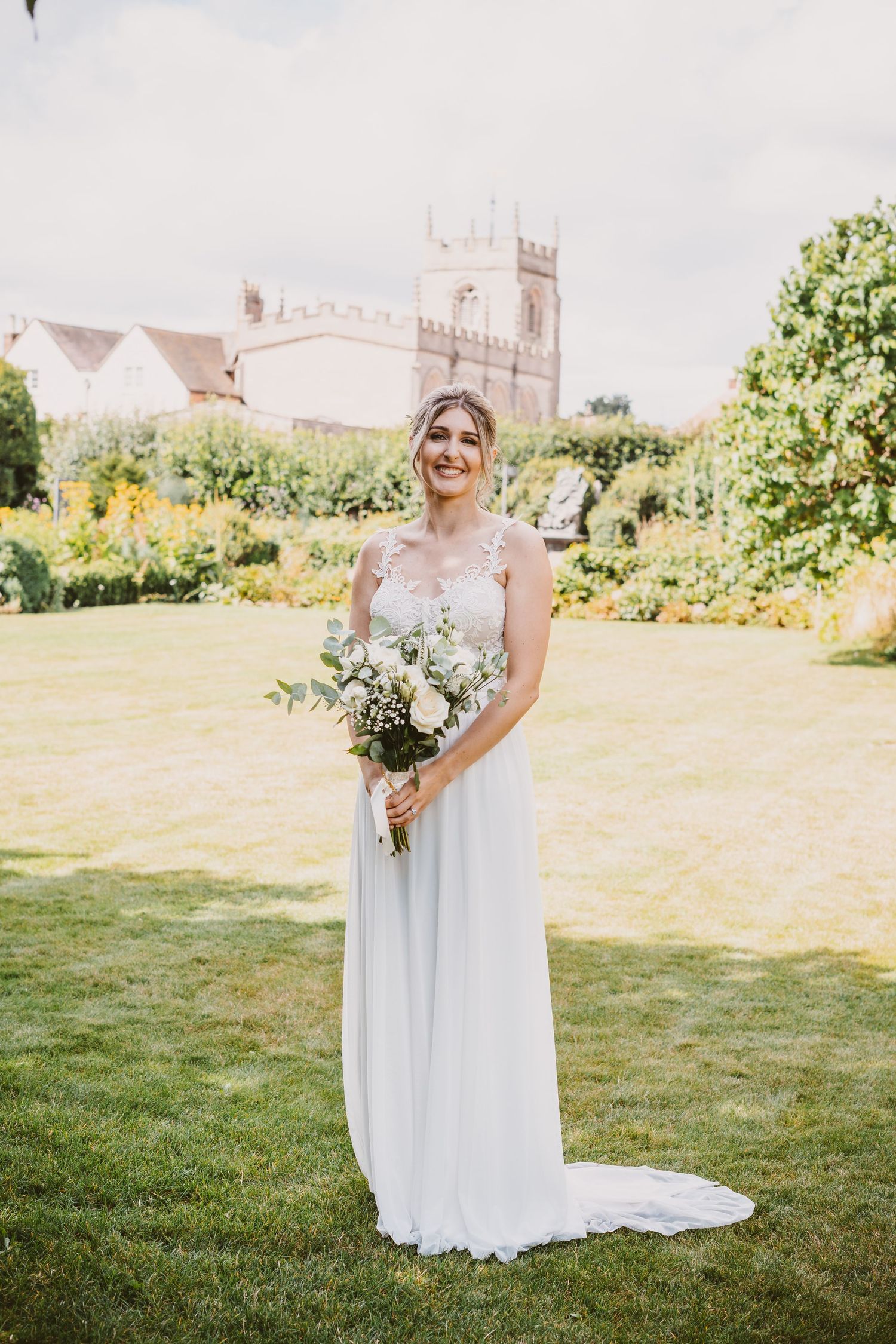 Elegant bride in flowing white dress holds bouquet in garden with historic building in background.