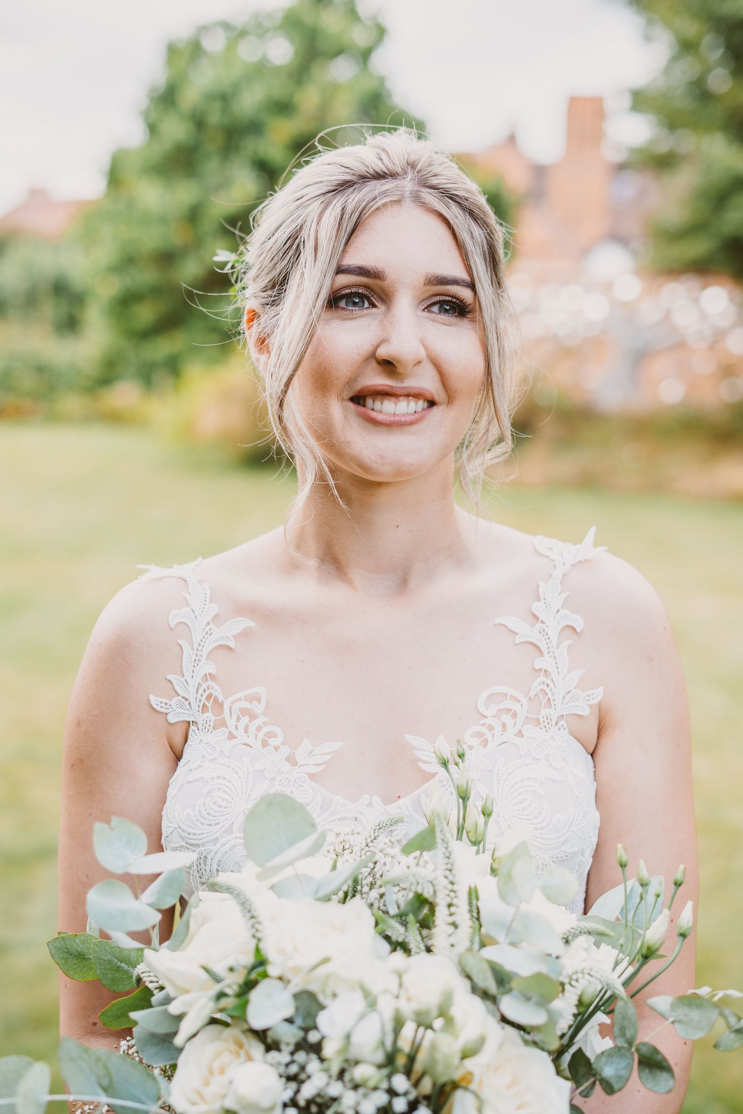 Bride in lace wedding dress smiles while holding white and green bouquet in garden setting.