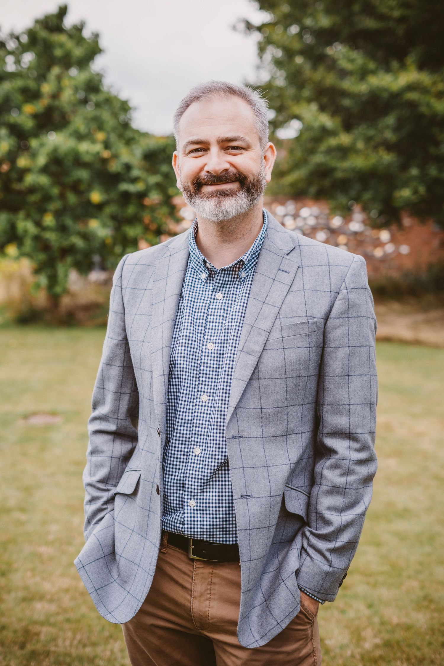 Groom in grey checkered suit jacket and blue gingham shirt stands in outdoor garden setting.