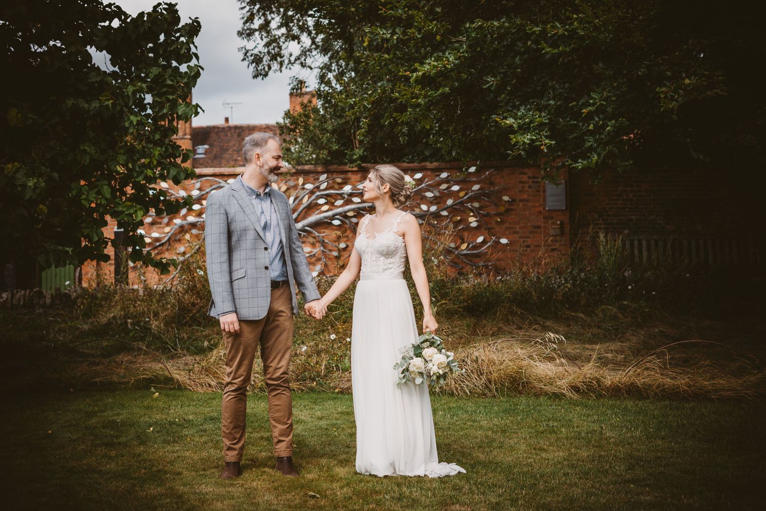 Wedding couple stands together in garden wearing grey suit and white lace dress.