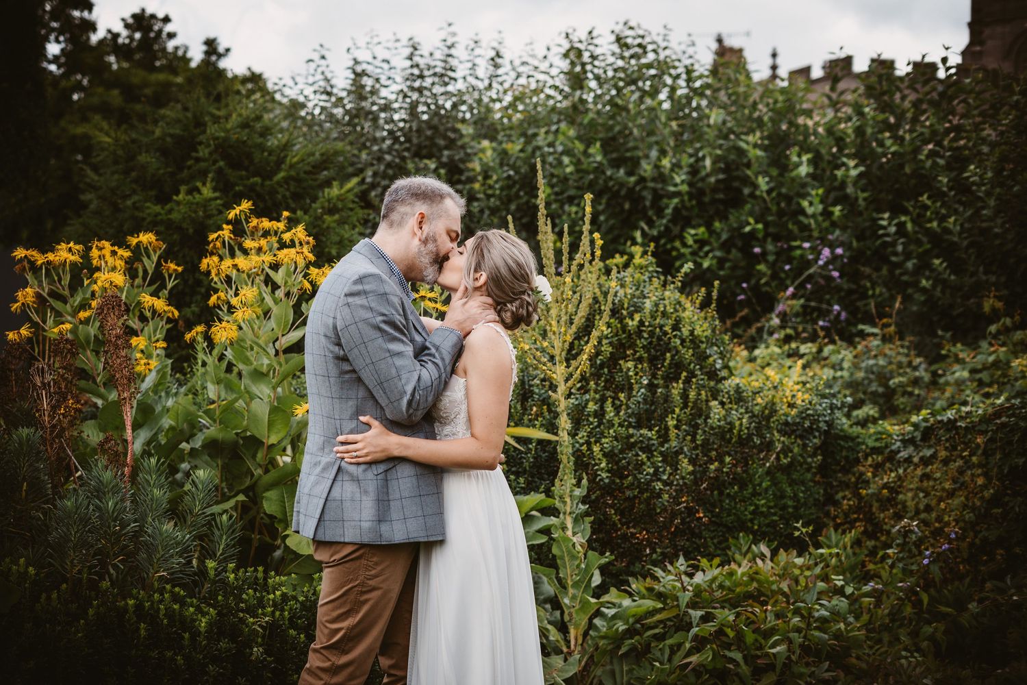 Romantic wedding couple embracing in garden with yellow flowers and greenery.
