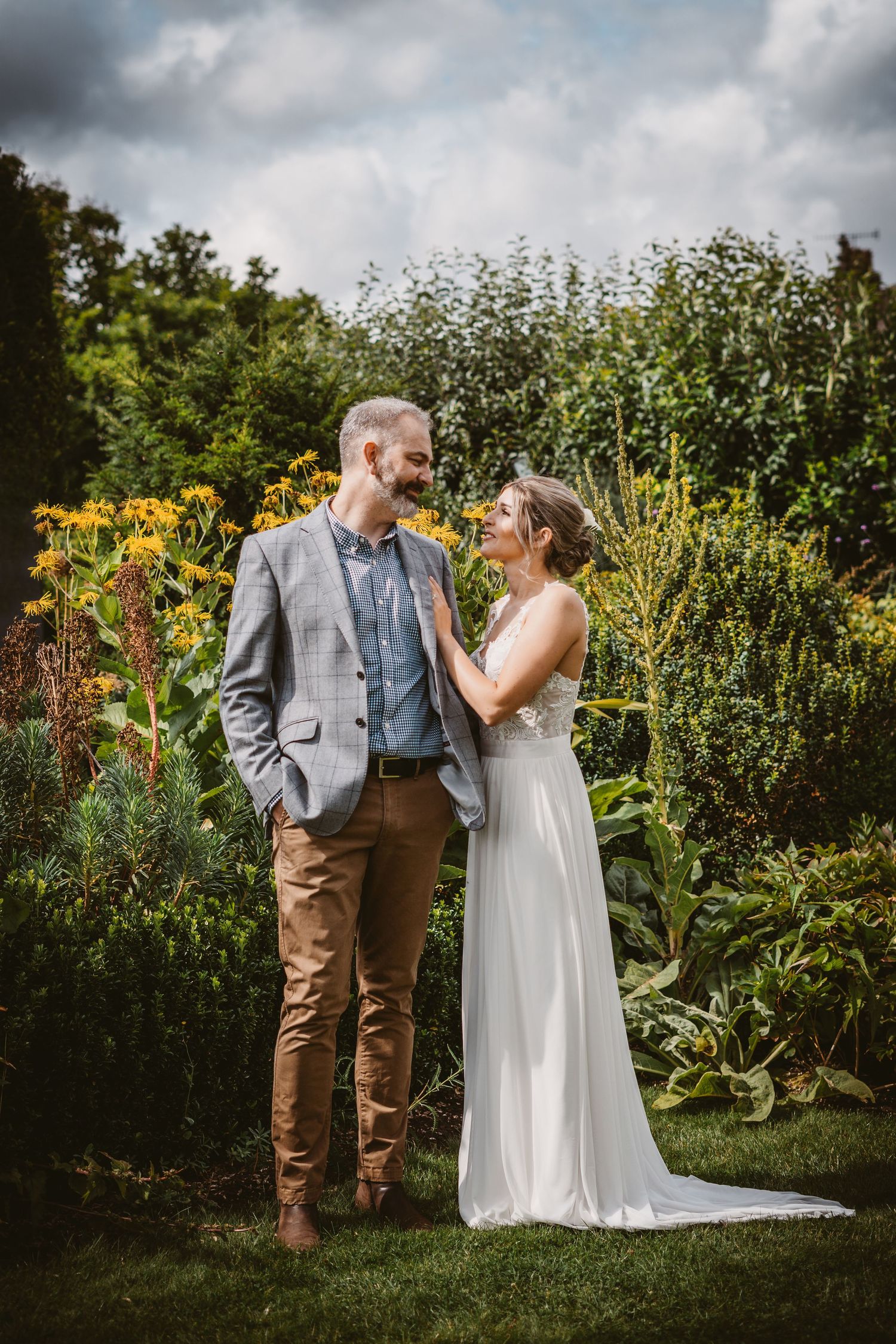 Wedding couple shares intimate moment in garden with yellow flowers and dramatic sky.