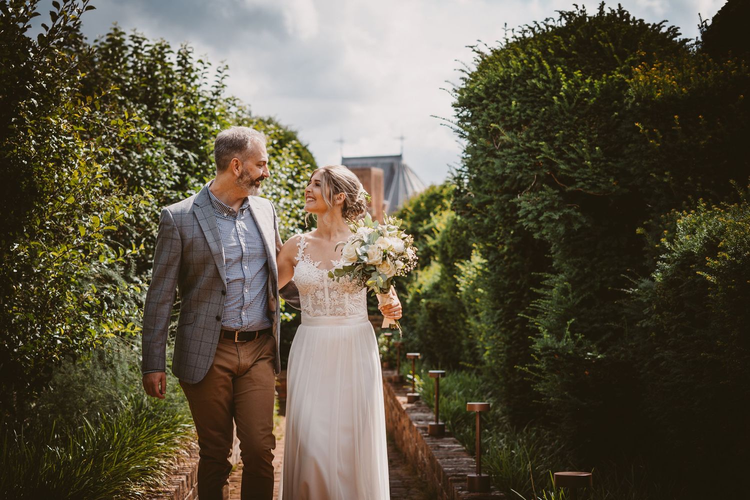 Newlyweds walking together on garden path surrounded by green hedges.