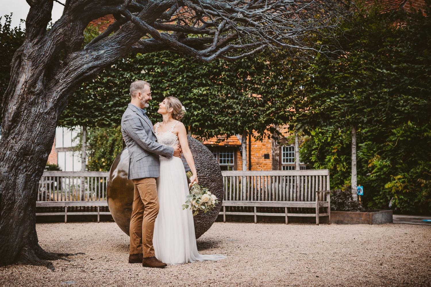 Wedding couple poses under large tree with wooden bench in background.
