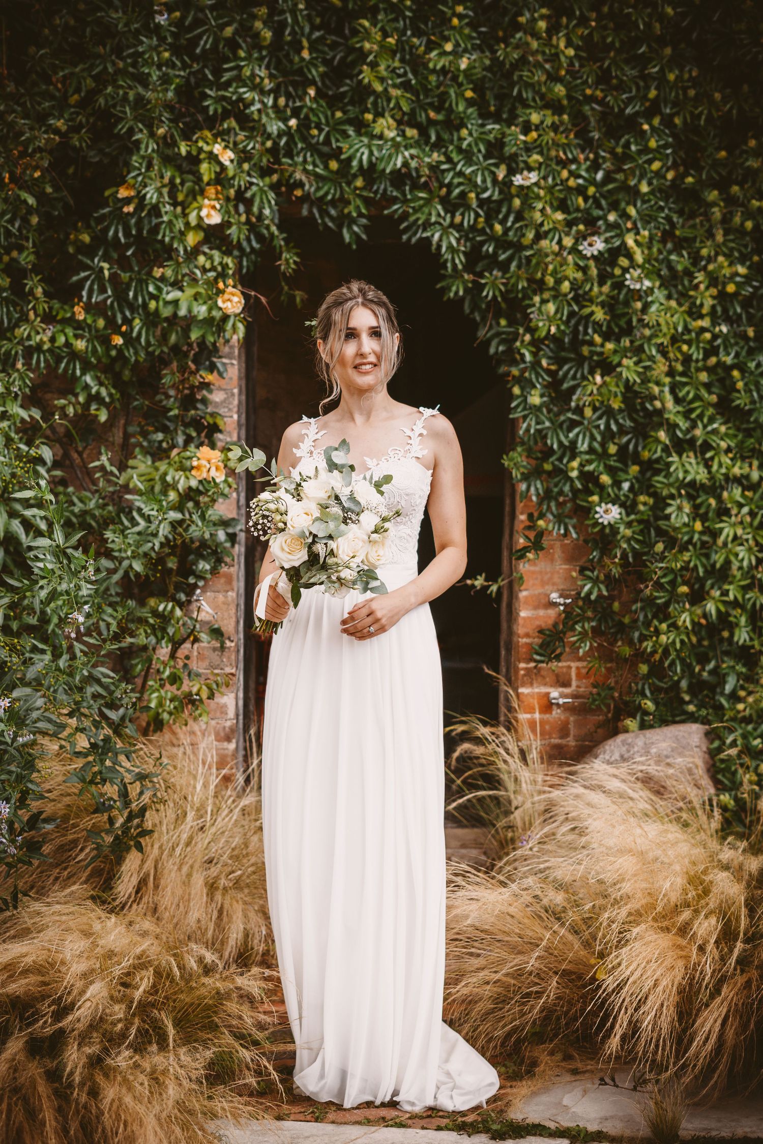 Bride in flowing white dress holds bouquet while standing in garden doorway.
