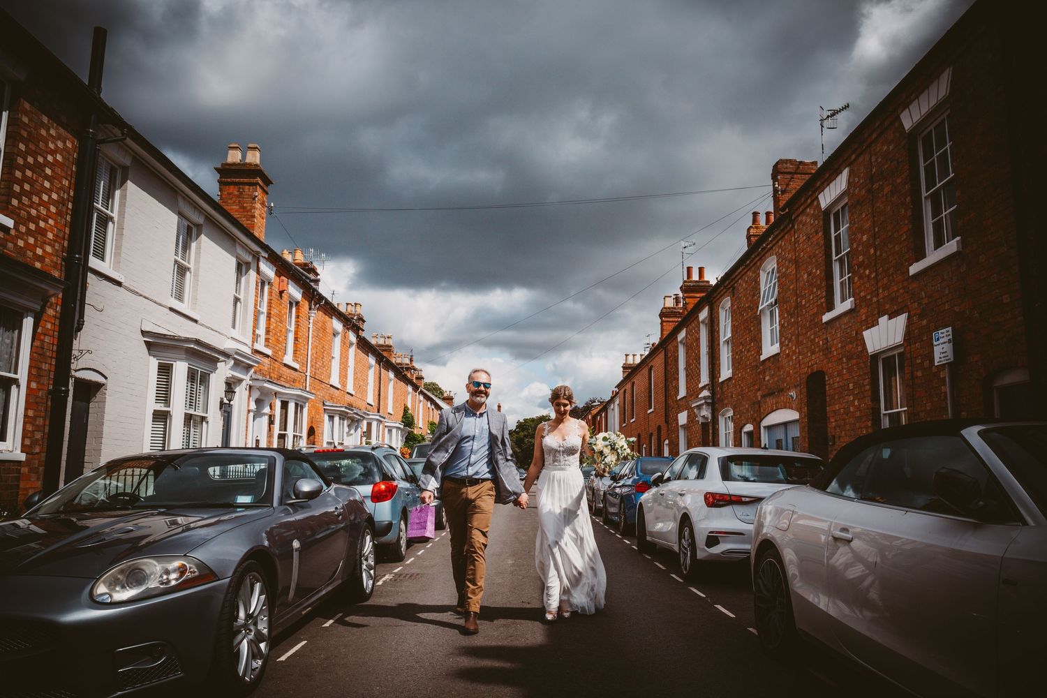 A wedding couple walking down a street lined with classic British terraced houses under stormy skies.
